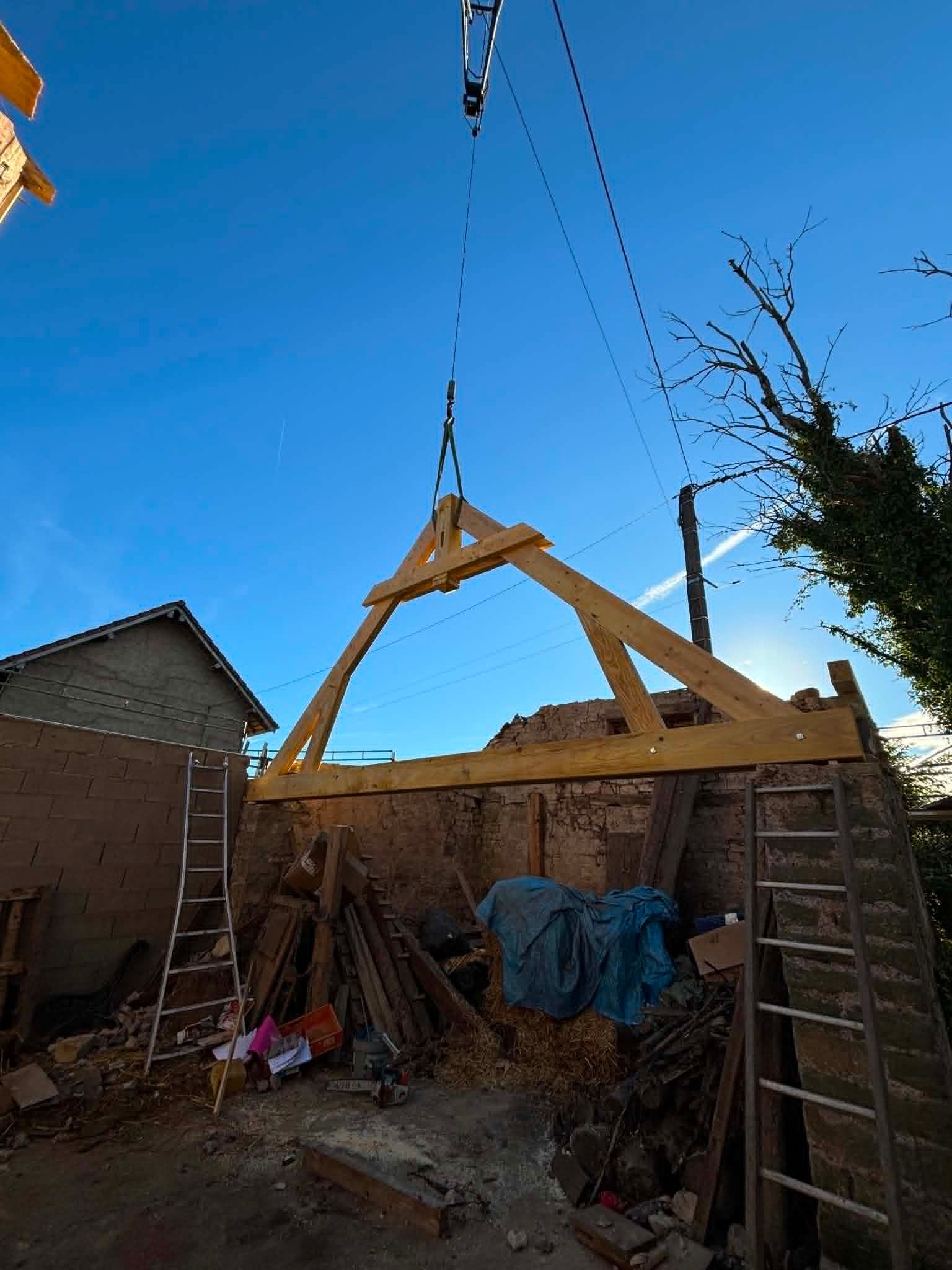 Sous un ciel bleu, une grue hisse une charpente de toit en bois au-dessus d'un chantier de construction dans une cour arrière.