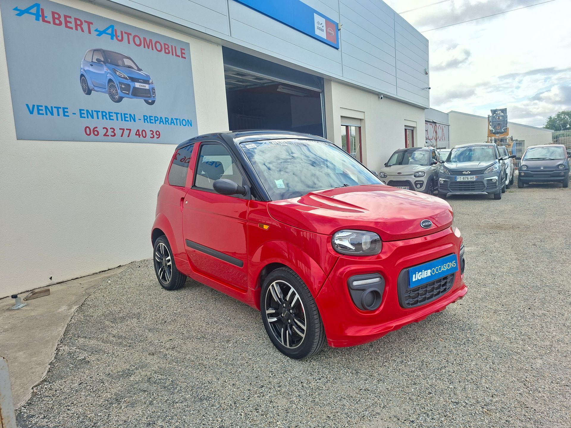 Voiture compacte rouge garée devant une concession automobile, avec d'autres véhicules sans permis en arrière-plan.