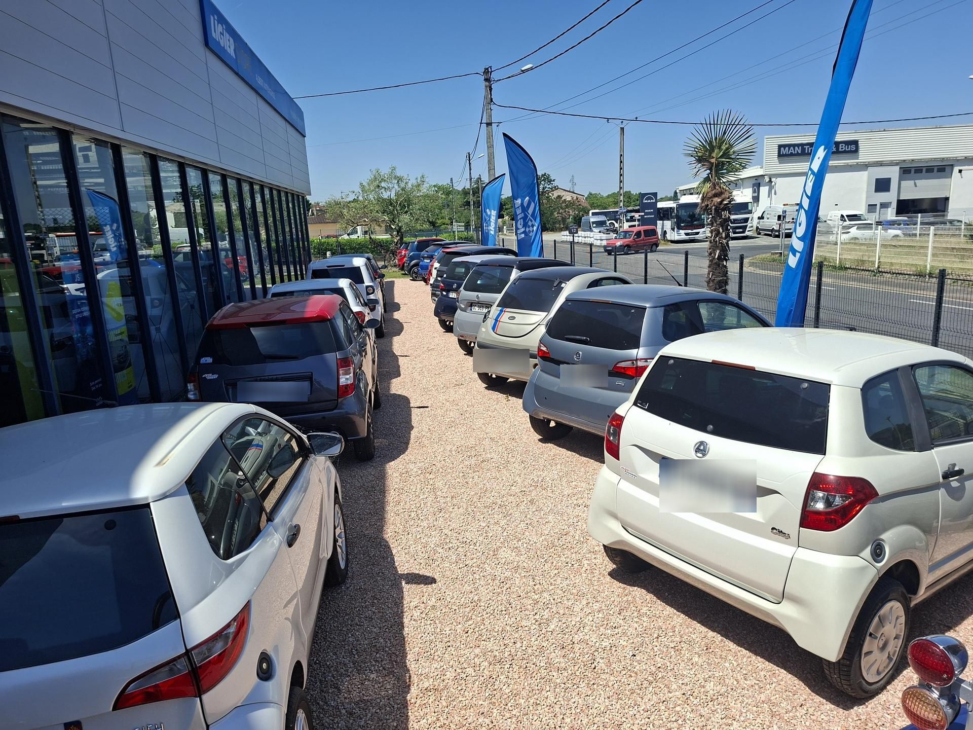 Parking d'un concessionnaire automobile avec des rangées de voitures garées à côté d'un bâtiment à façade vitrée par une journée ensoleillée.