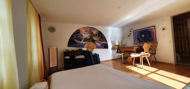 Schlafzimmer mit Blick auf See und Berge durch eine Balkontür. Zwei Fenster mit roten Vorhängen. Weißes Bett.