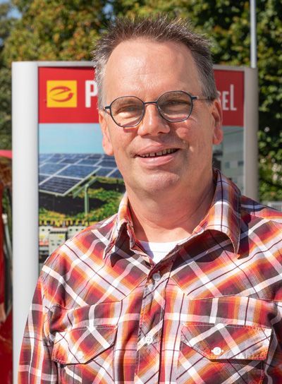 Ein Mann mit Brille und kariertem Hemd lächelt vor einer Solarpanel-Anzeigetafel.