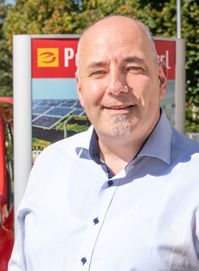 Ein Mann in blauem Hemd lächelt vor einem roten Schild mit Solarzellen.