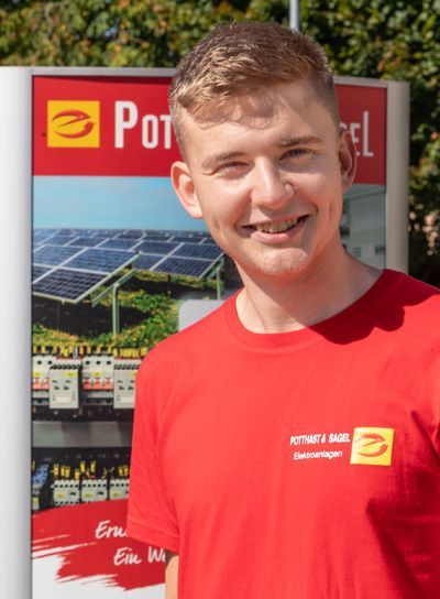 Mann in rotem Hemd lächelt vor einem Schild mit Solarzellen.