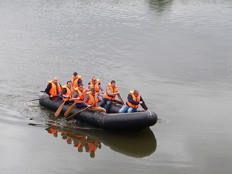 Menschen in orangefarbenen Schwimmwesten rudern ein schwarzes Schlauchboot auf einem See.