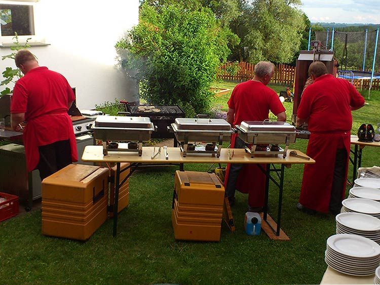 Drei Personen in roten Uniformen bereiten Speisen an einem Buffet im Freien mit Warmhaltebehältern und Grillgeräten zu.