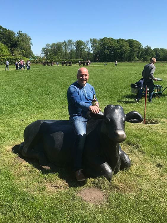 Mann sitzt an einem sonnigen Tag auf einer schwarzen Kuhstatue auf einer grasbewachsenen Wiese.