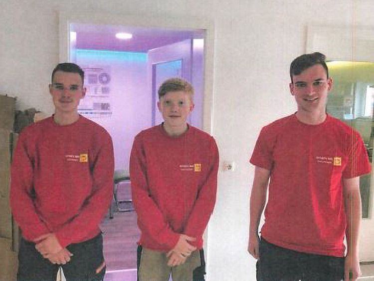 Drei Personen in roten Hemden stehen in einem Innenraum. Im Hintergrund sind ein Türrahmen und ein Teil einer Theke zu sehen.