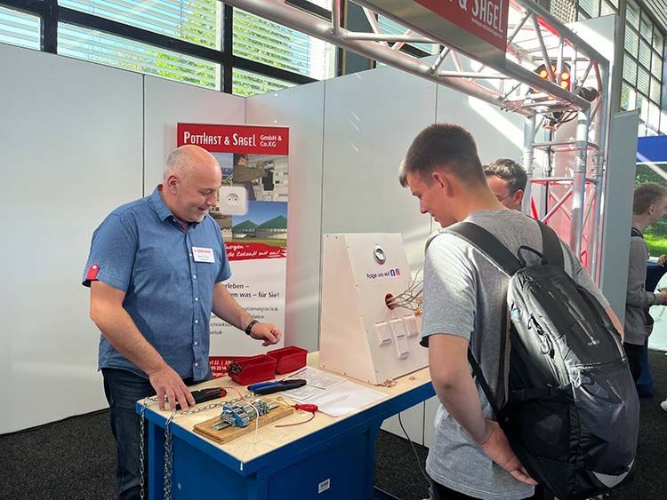Ein Mann demonstriert elektrische Bauteile an einem Messestand; zwei Männer beobachten ihn; Messeatmosphäre.