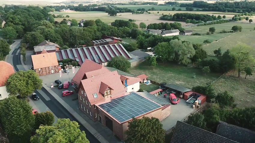 Luftaufnahme eines Bauernhofs mit rotgedeckten Gebäuden, Solarpaneelen und umliegenden grünen Feldern.