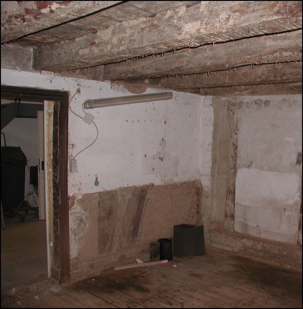 Verfallenes Zimmerinneres mit freiliegenden Balken, abgeplatzten Wänden und einer Türöffnung.
