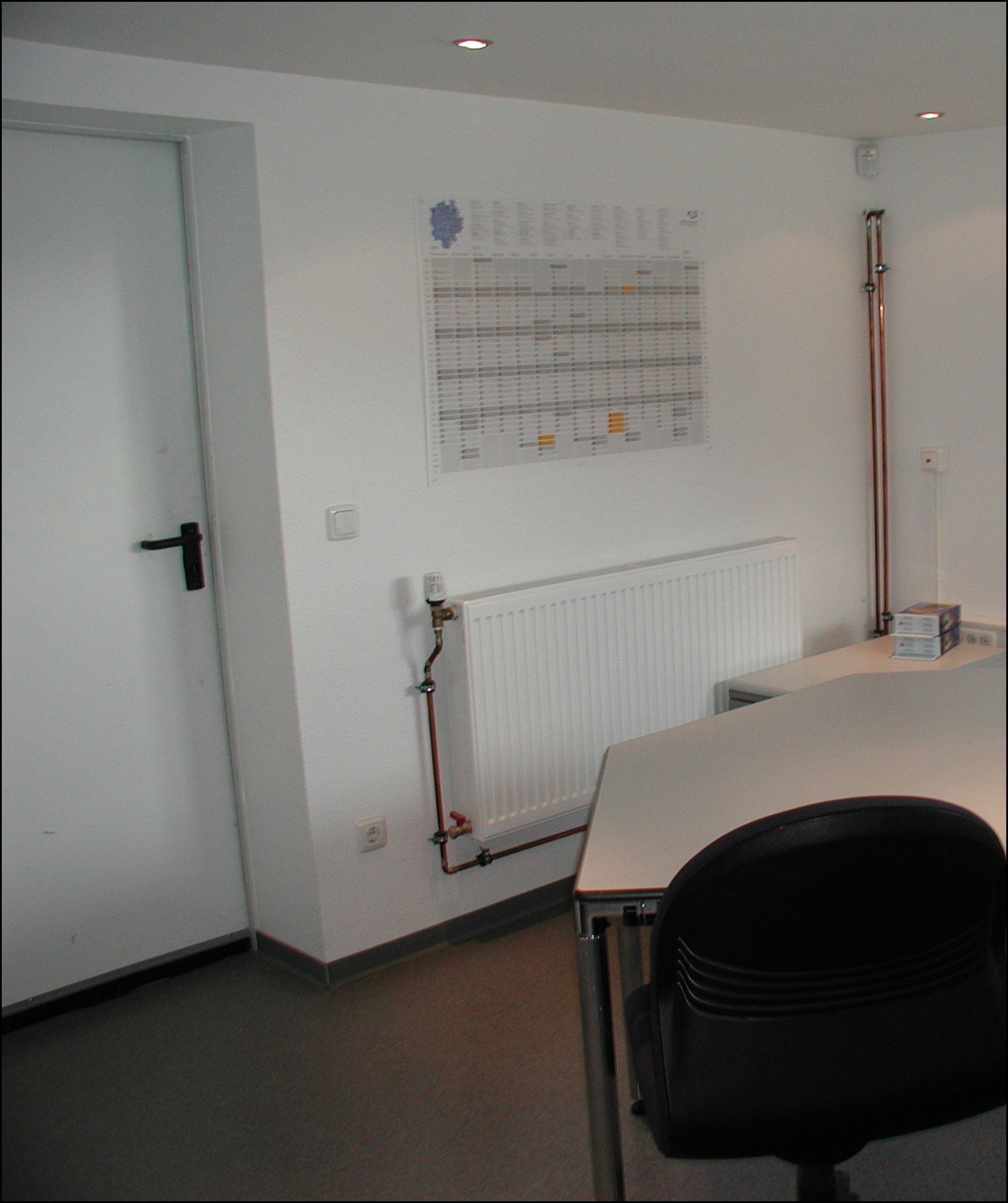 Weißes Büro mit geschlossener Tür, Heizkörper, Schreibtisch, Stuhl und einem Bauplan an der Wand.