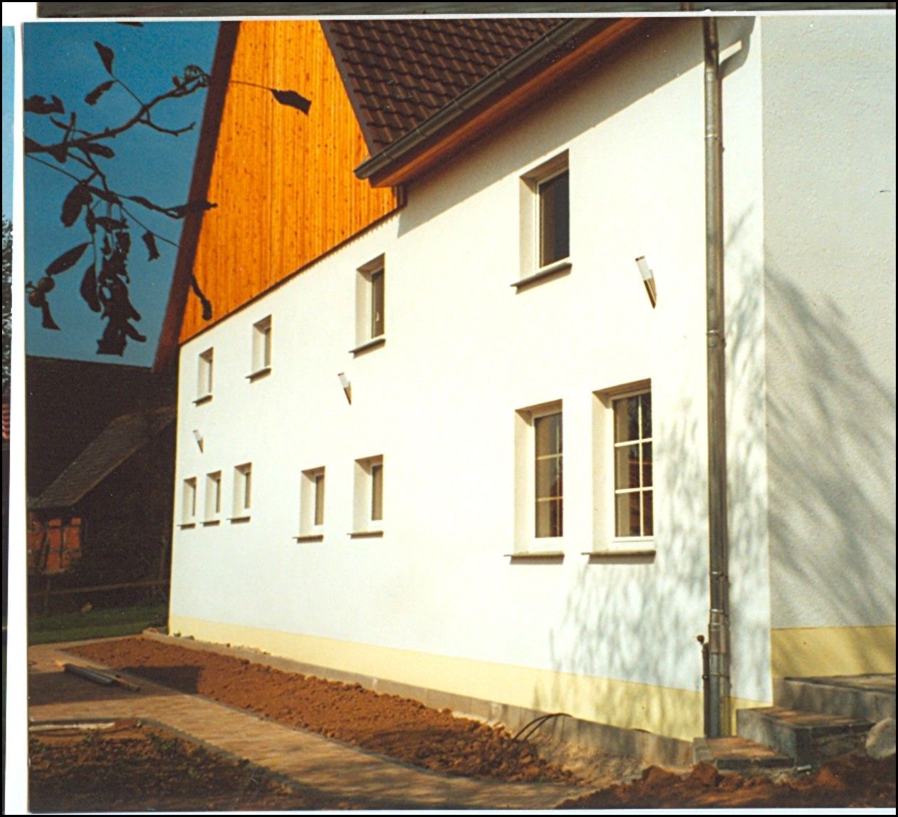 Weißes Gebäude mit Holzdach, Fenstern und Fallrohr. Brauner Boden und Stufen im Vordergrund.
