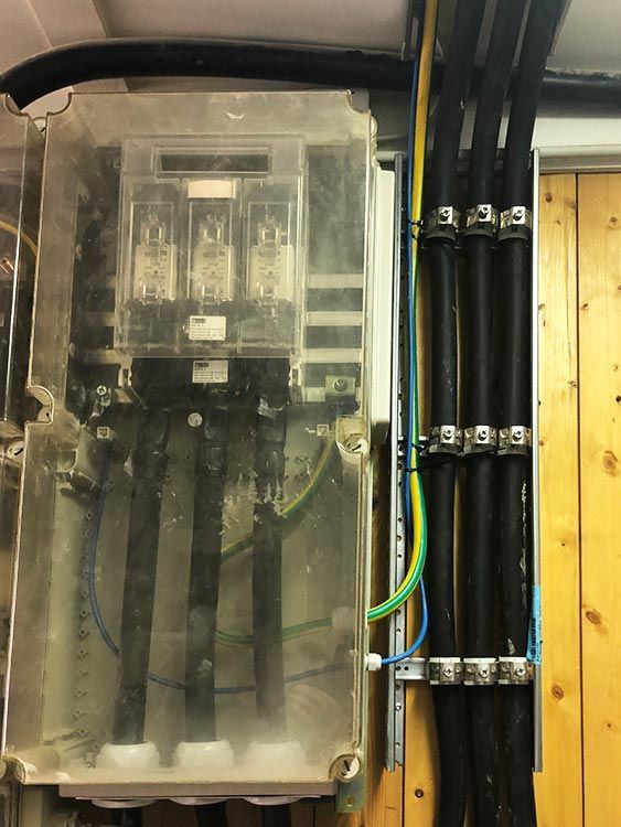 Elektrischer Verteilerkasten mit drei Sicherungen, dicken schwarzen Kabeln und Erdungsdrähten. An einer Holzwand montiert.