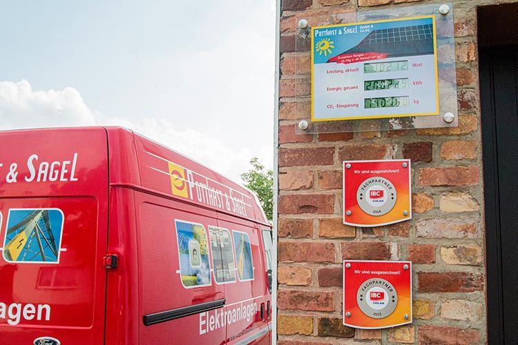 Ein roter Lieferwagen neben einer Mauer mit Überwachungsdaten für Solarpaneele und zwei Ladestationen für Elektrofahrzeuge.