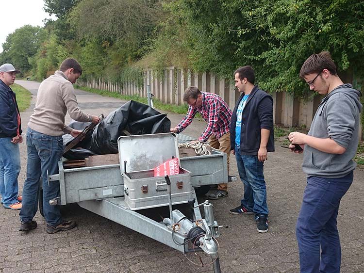 Männer verladen im Freien Gegenstände in einen Anhänger. Einer der Männer trägt eine Brille, die anderen sind leger gekleidet