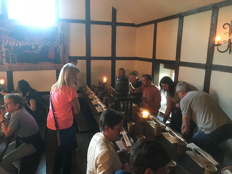 Menschen in einem langen Raum mit Holztischen, einige stehen, einige sitzen, Kerzen brennen und ein Fass steht daneben.