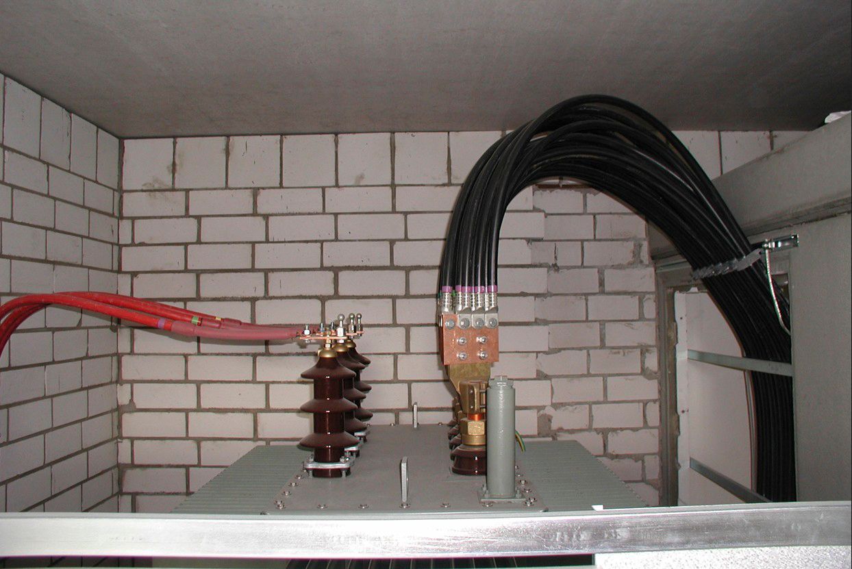 Elektrischer Transformator mit Kabeln in einem gemauerten Raum. Schwarze und rote Drähte sind an das Gerät angeschlossen.