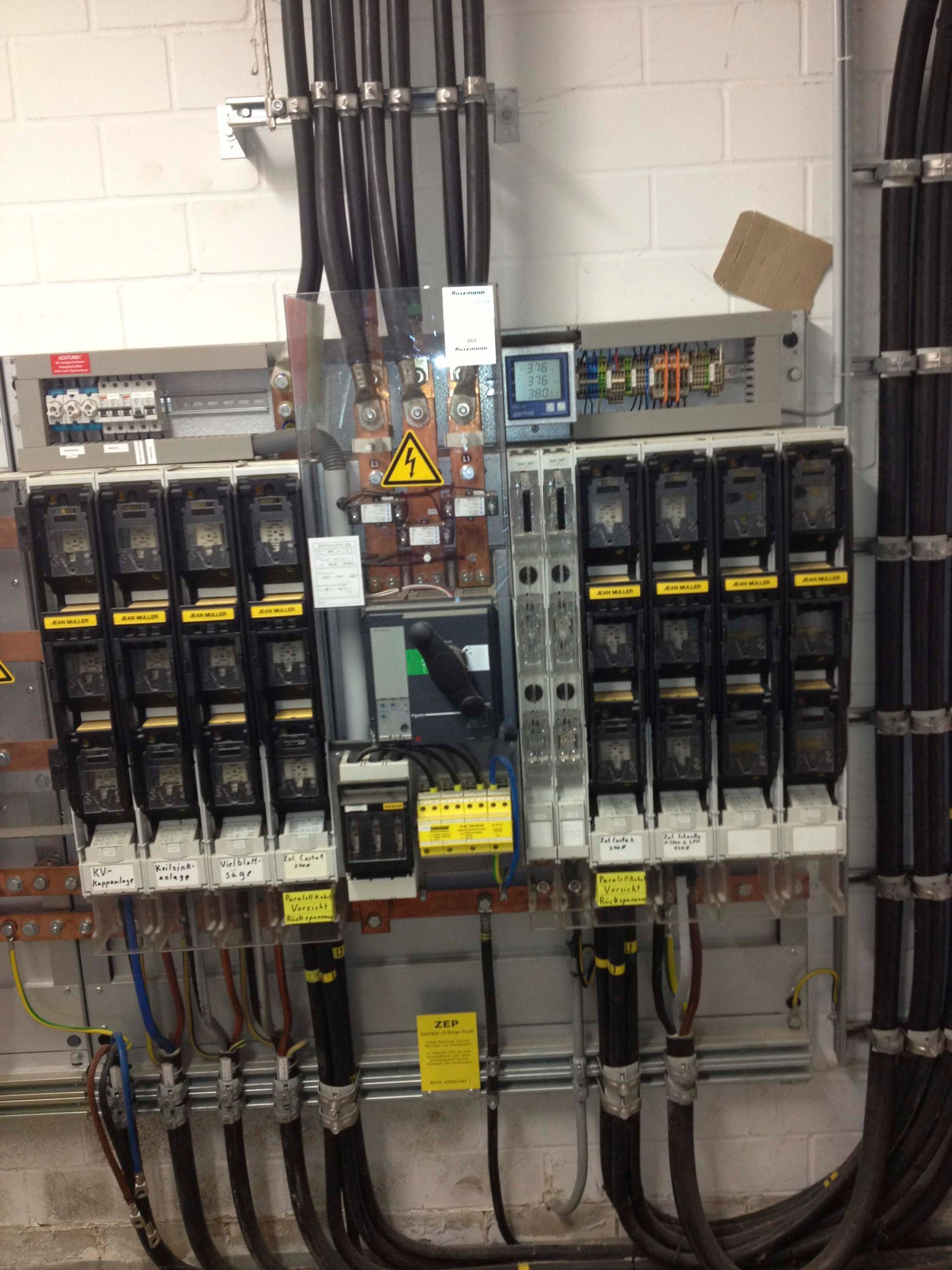 Elektrischer Verteilerkasten mit zahlreichen schwarzen Kabeln, Schaltern und Beschriftungen an einer weißen Backsteinwand.