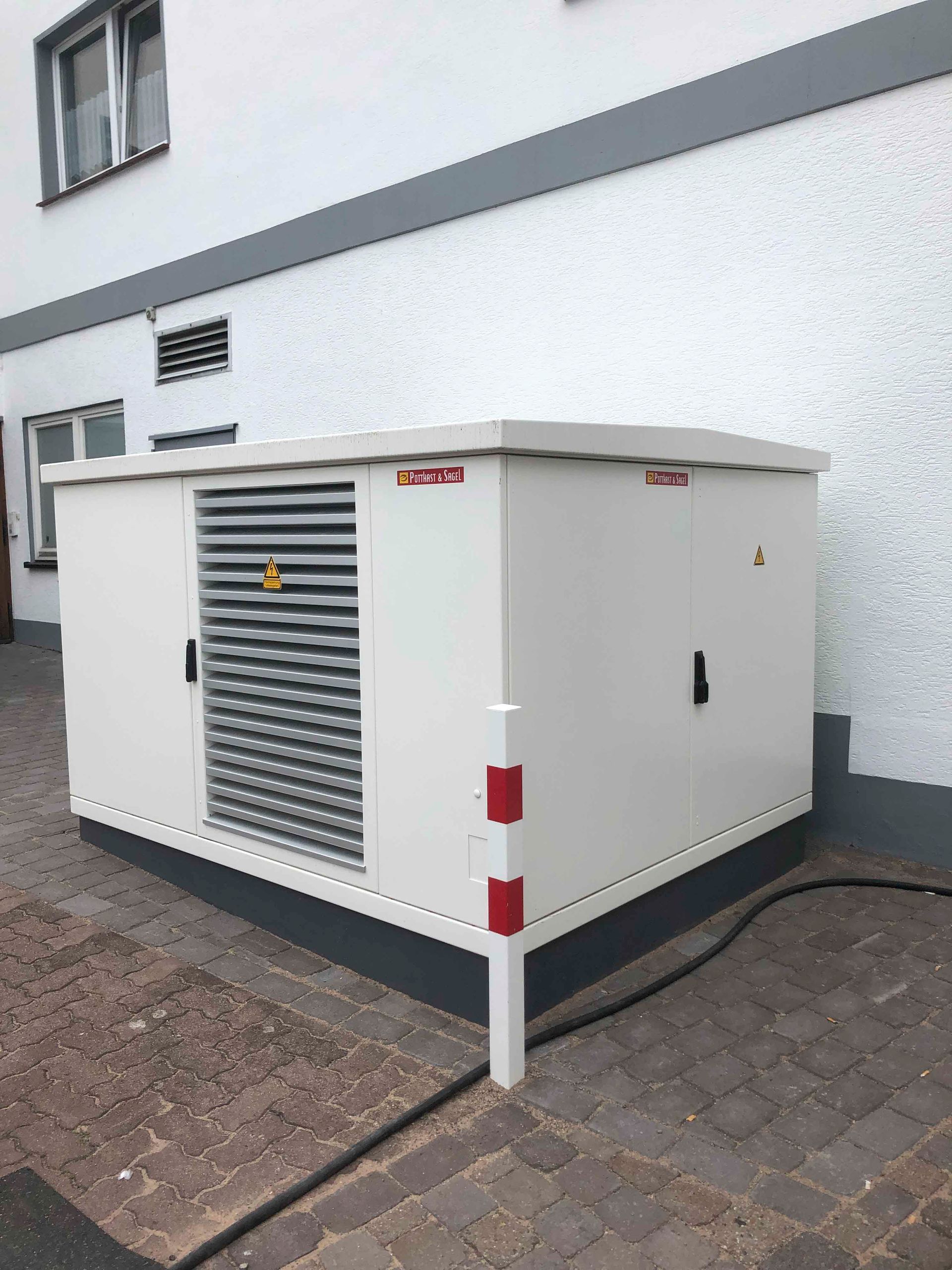 Weißer Elektroverteilerkasten mit grauem Lüftungsschlitz an einem Gebäude auf einer Ziegelsteinfassade.