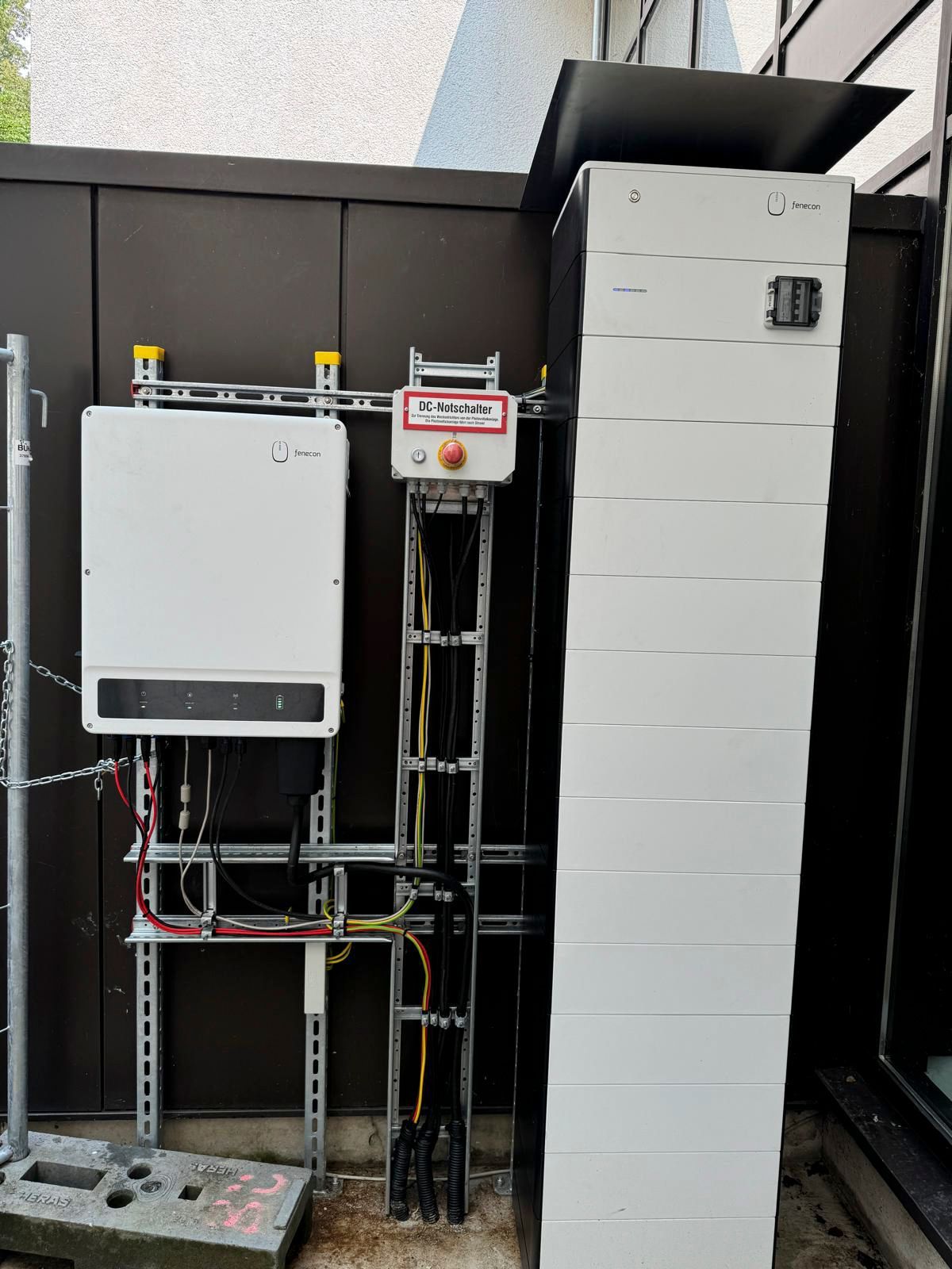 Komponenten für die Außensolaranlage, die an einer Wand montiert sind. Weißer Wechselrichter, ein Batterieturm und Kabel