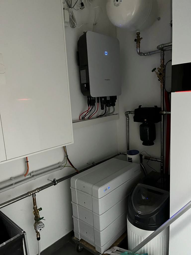 Solarwechselrichter und Batteriesystem im Hauswirtschaftsraum installiert. Weiße und graue Haushaltsgeräte vor weißer Wand.