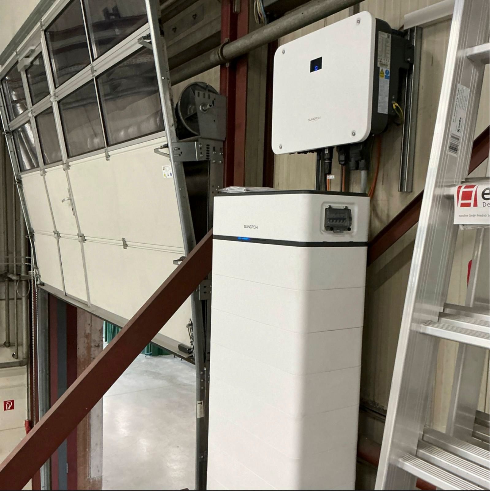 Weiße Solaranlage neben weißer Batterie, an der Wand einer Garage montiert. Rechts befindet sich eine Leiter.