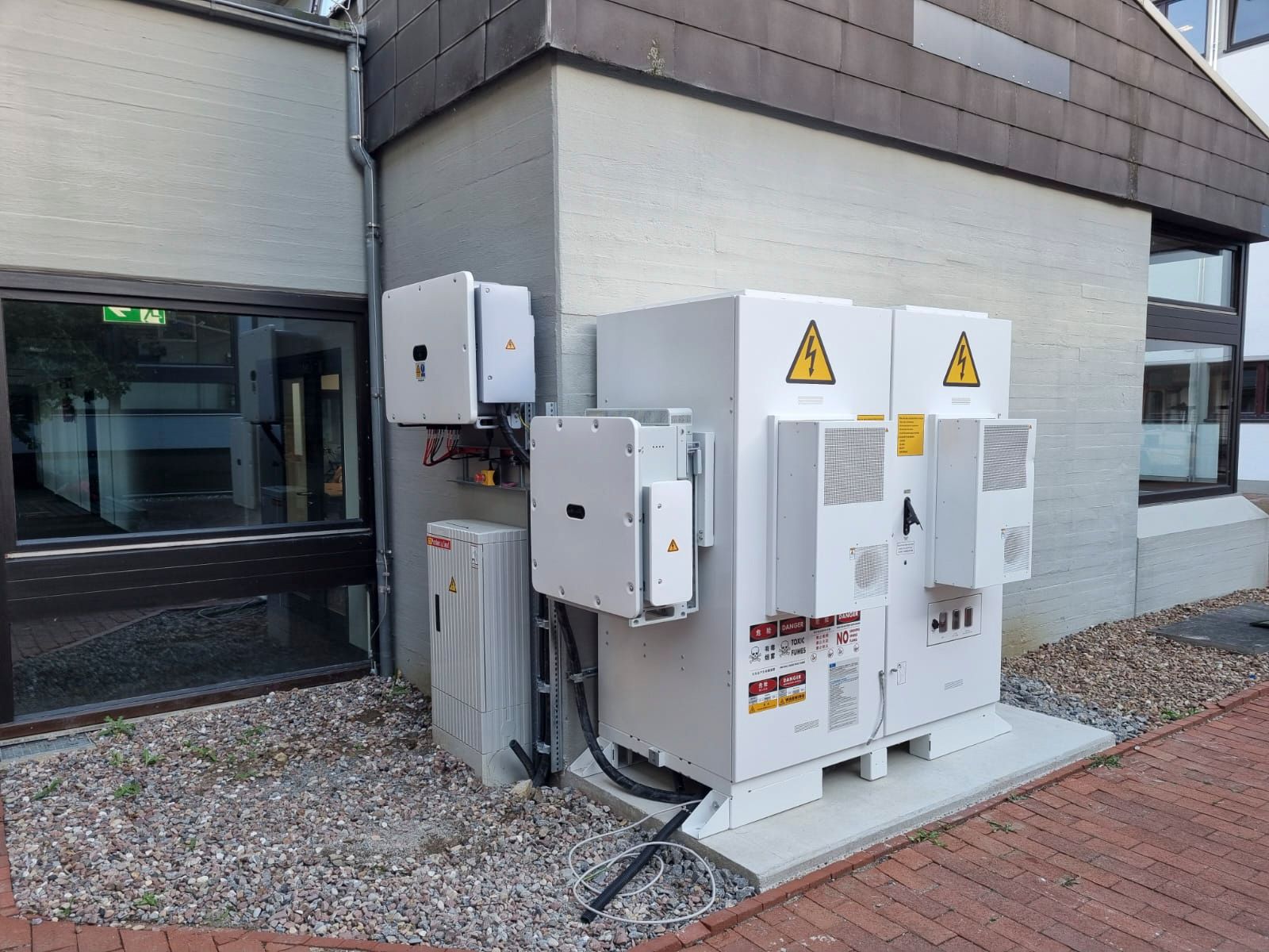 Außenansicht von elektrischen Anlagen, vermutlich für Solarpaneele, an einer Gebäudeecke. Weiße Schränke mit Warnsymbolen.