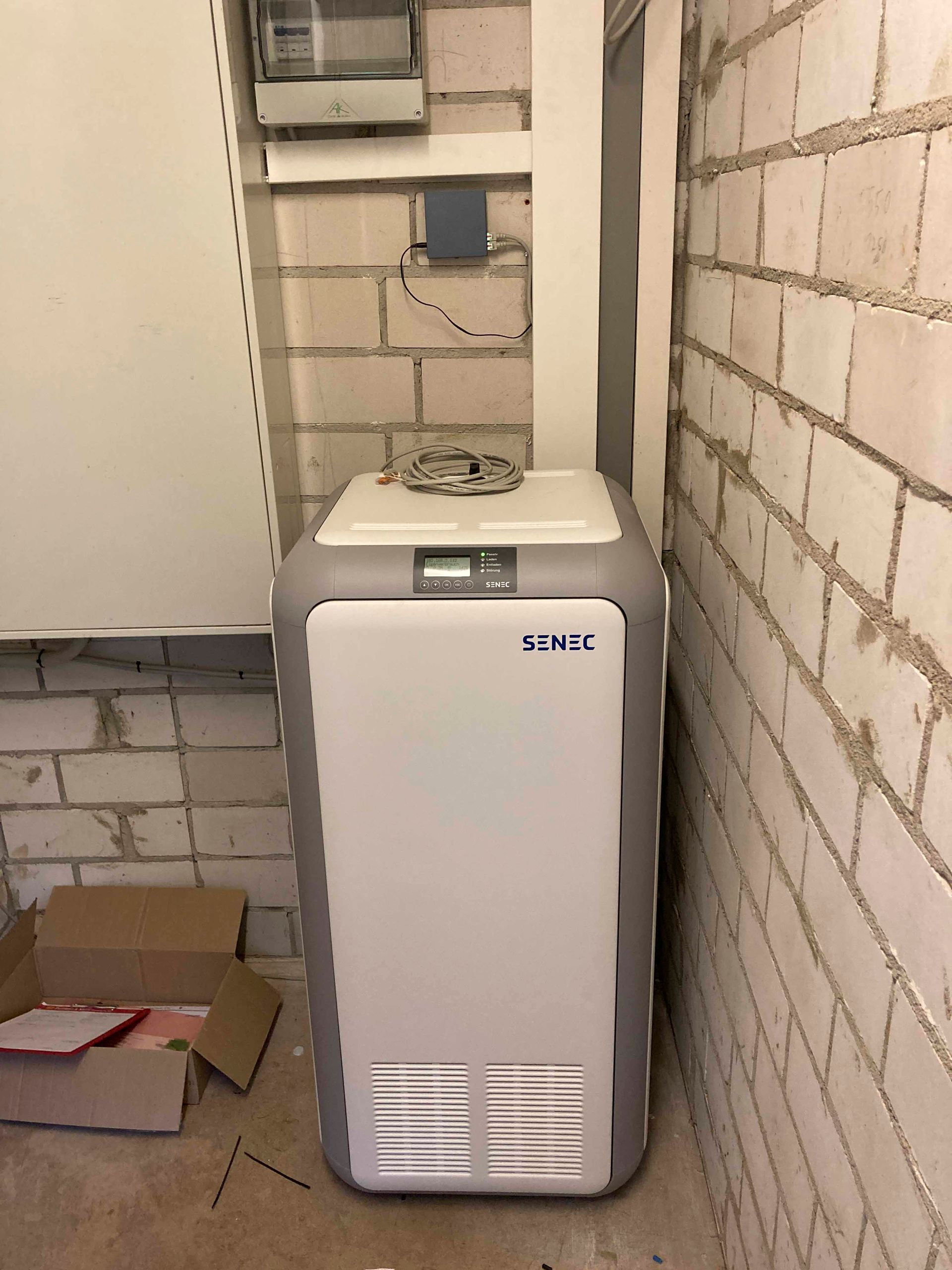 Große, weiß-graue Maschine vor einer Backsteinmauer, vermutlich Laborausrüstung.