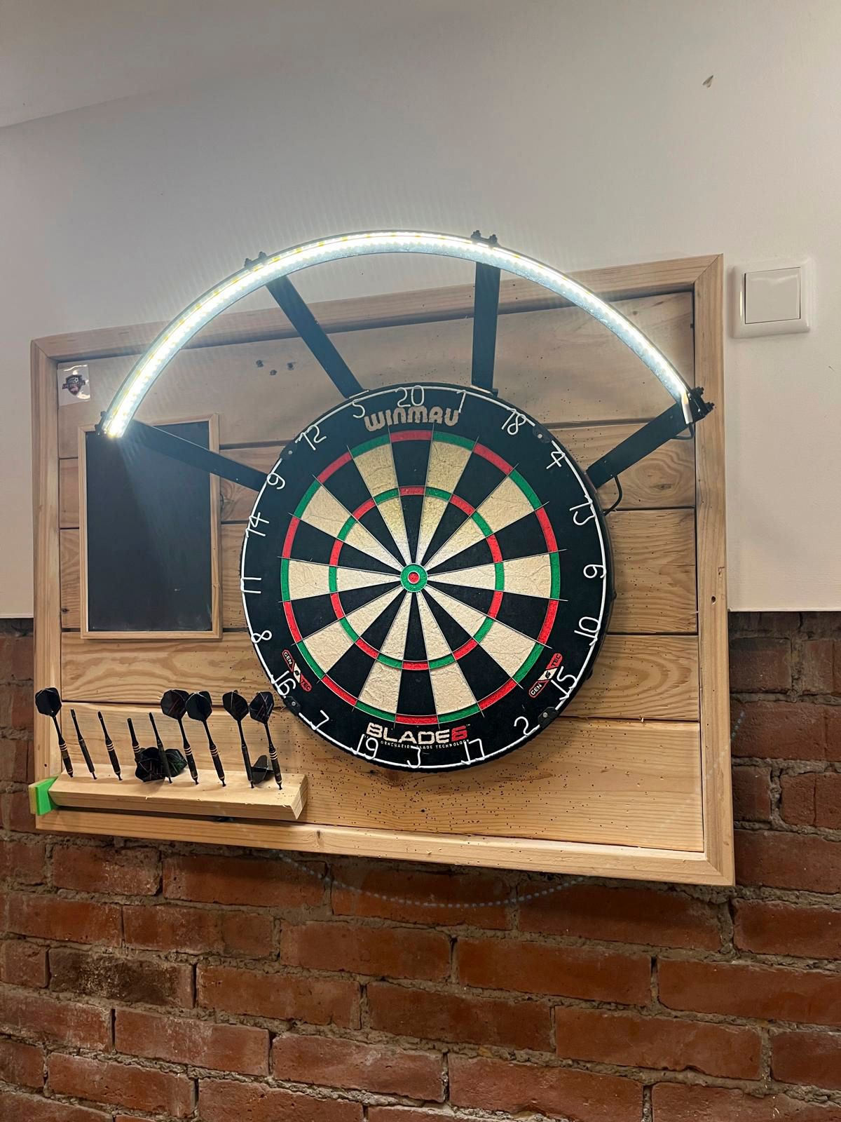 Dartscheibenaufbau mit Deckenbeleuchtung und Dartpfeilhaltern an einer Backsteinwand.