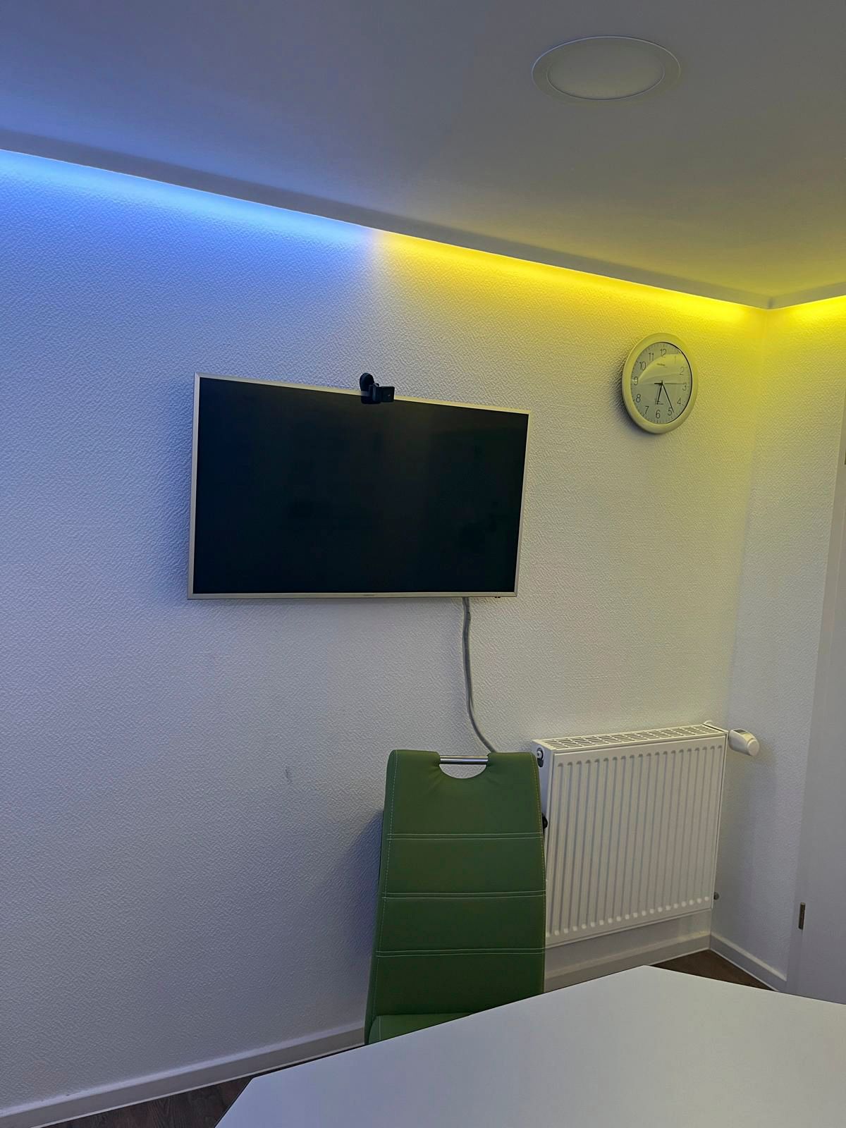 Fernseher an weißer Wand mit blau/gelber LED-Beleuchtung. Grüner Stuhl und weißer Heizkörper sind zu sehen.