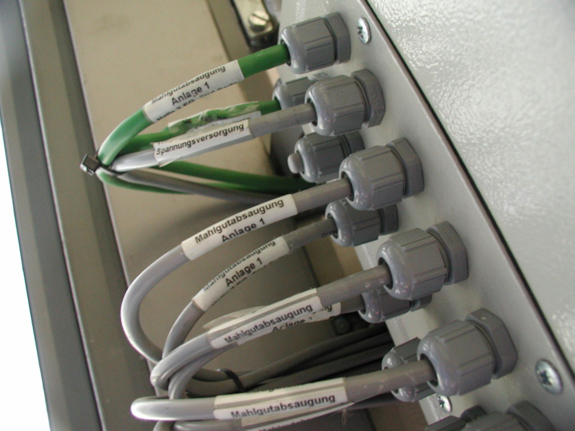 Nahaufnahme von beschrifteten grauen Kabeln, die durch graue Kabelverschraubungen in eine Schalttafel geführt werden