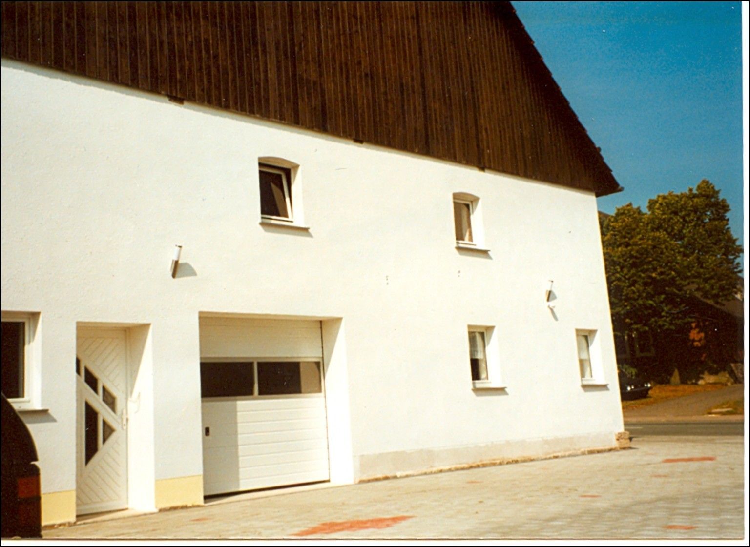 Weißes Gebäude mit braunem Dach, mehreren kleinen Fenstern und einem Garagentor.
