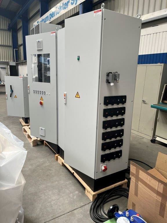 Graue Elektroschränke auf einer Holzpalette in einer Werkstatt. Verschiedene Bedienelemente, Beschriftungen und Verkabelung