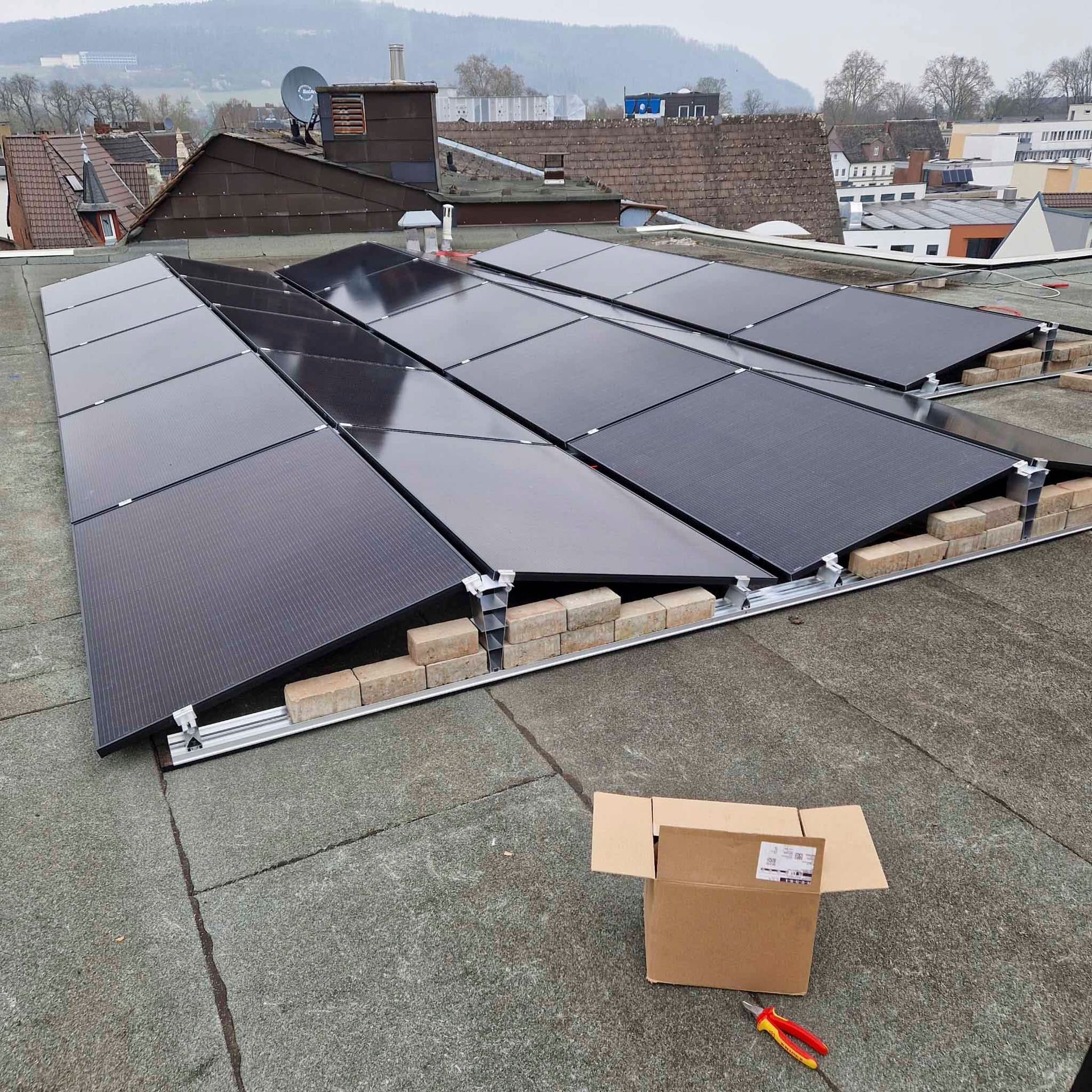 Auf einem Flachdach sind Solarpaneele installiert, daneben stehen ein Karton und Werkzeug.
