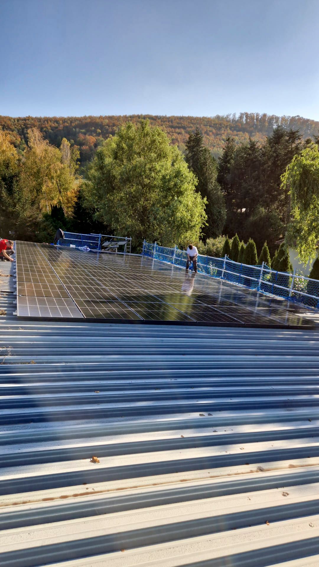 Auf einem Wellblechdach installierte Solarpaneele, im Hintergrund Bäume und ein Hügel.