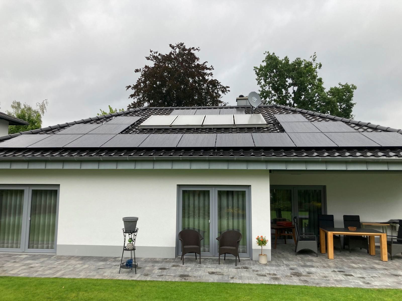 Haus mit Solarpaneelen auf dem Dach, hellgraue Fassade, Terrasse mit Gartenmöbeln, bewölkter Himmel.