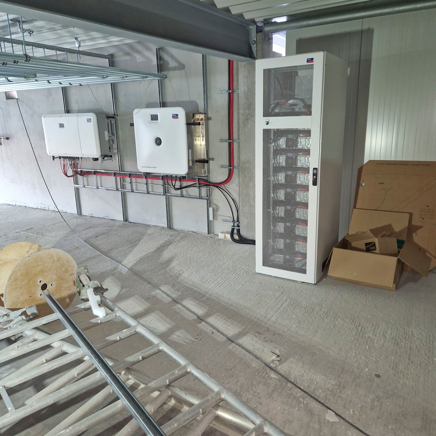 Innenansicht eines Technikraums mit Solarwechselrichtern und einem großen Serverschrank. Betonboden, graue Wände.