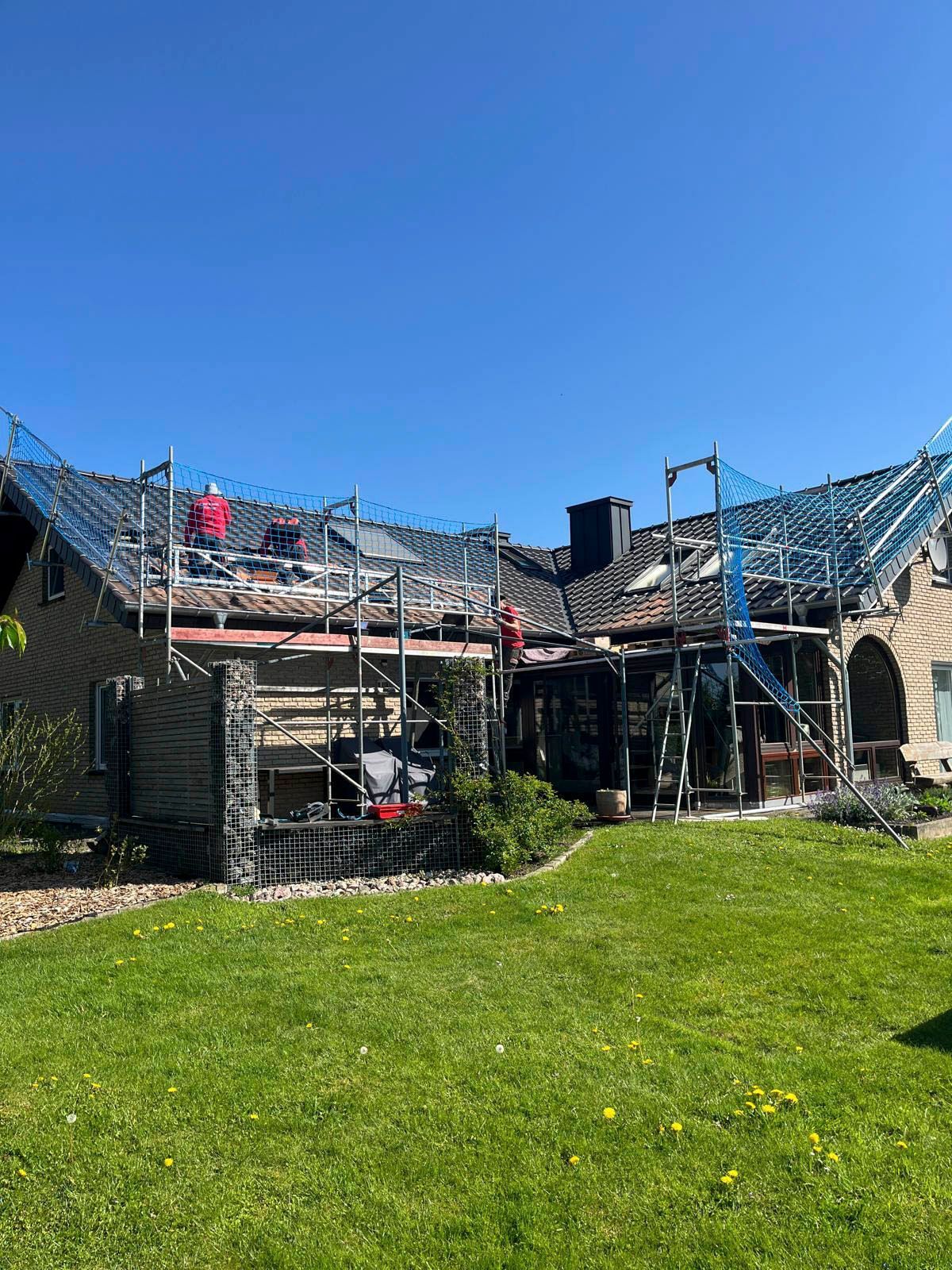 Das Hausdach wird gerade eingebaut, Gerüst und Sicherheitsnetze sind angebracht. Grüner Rasen und blauer Himmel.