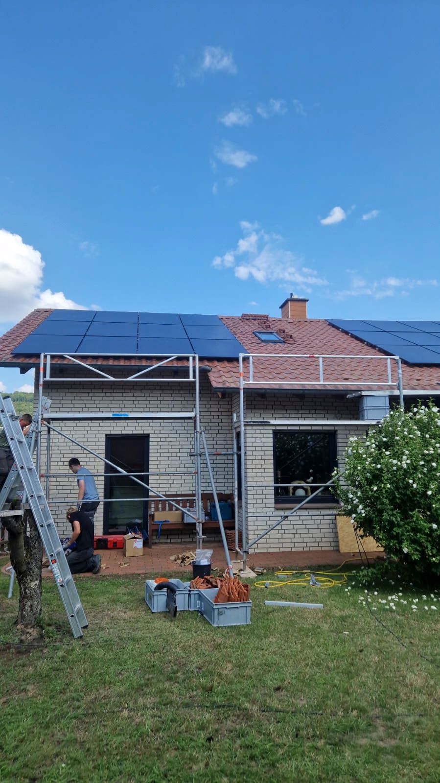 Haus mit auf dem Dach installierten Solarpaneelen; Arbeiter auf einem Gerüst, blauer Himmel.