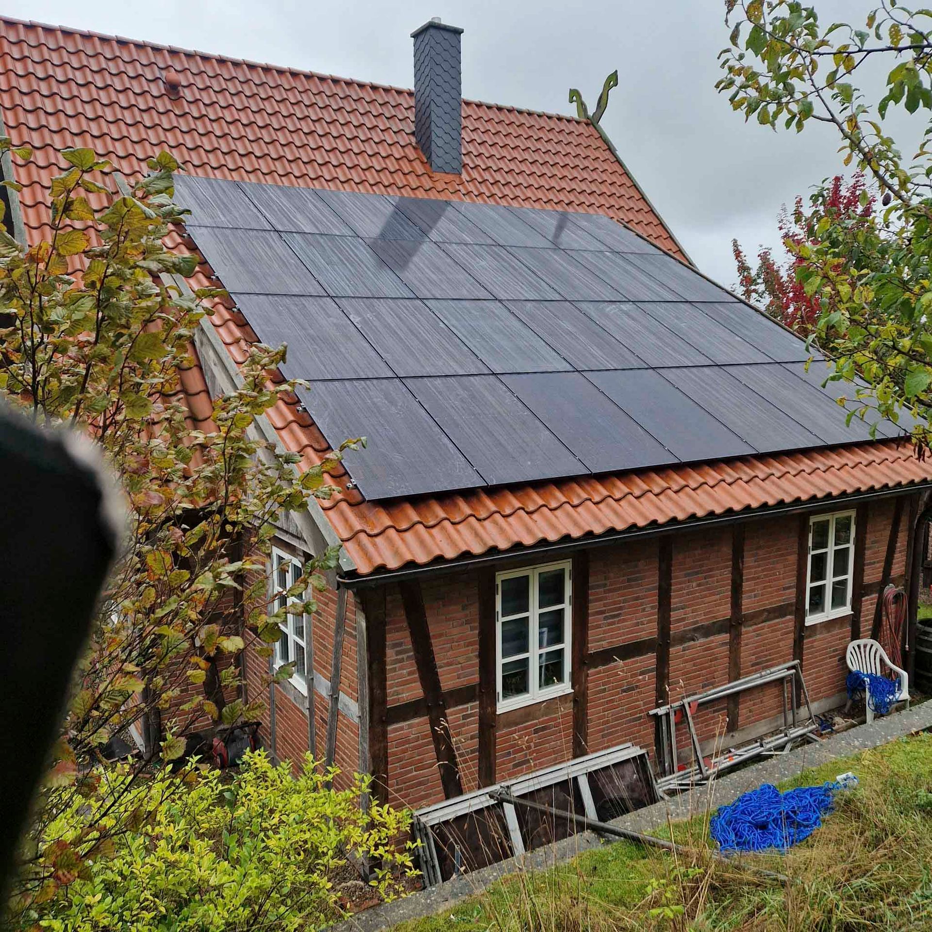 Solarpaneele auf dem rotziegelgedeckten Dach eines Backsteinfachwerkhauses mit weißgerahmten Fenstern, umgeben von Bäumen.