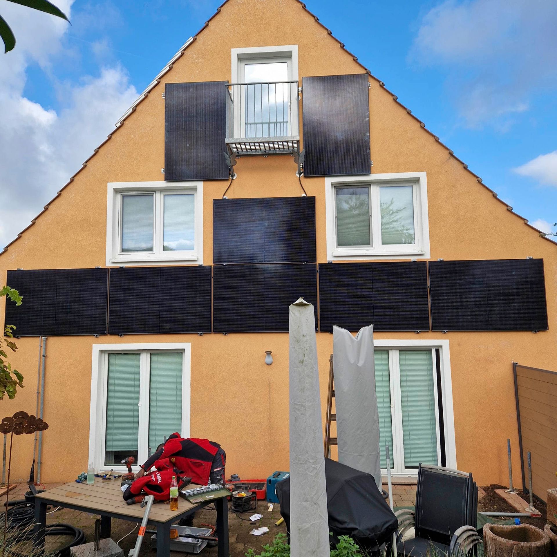 Gelbes Haus mit an der Fassade angebrachten Solarpaneelen. Fenster und Balkon sind sichtbar. Baumaschinen im Vordergrund.
