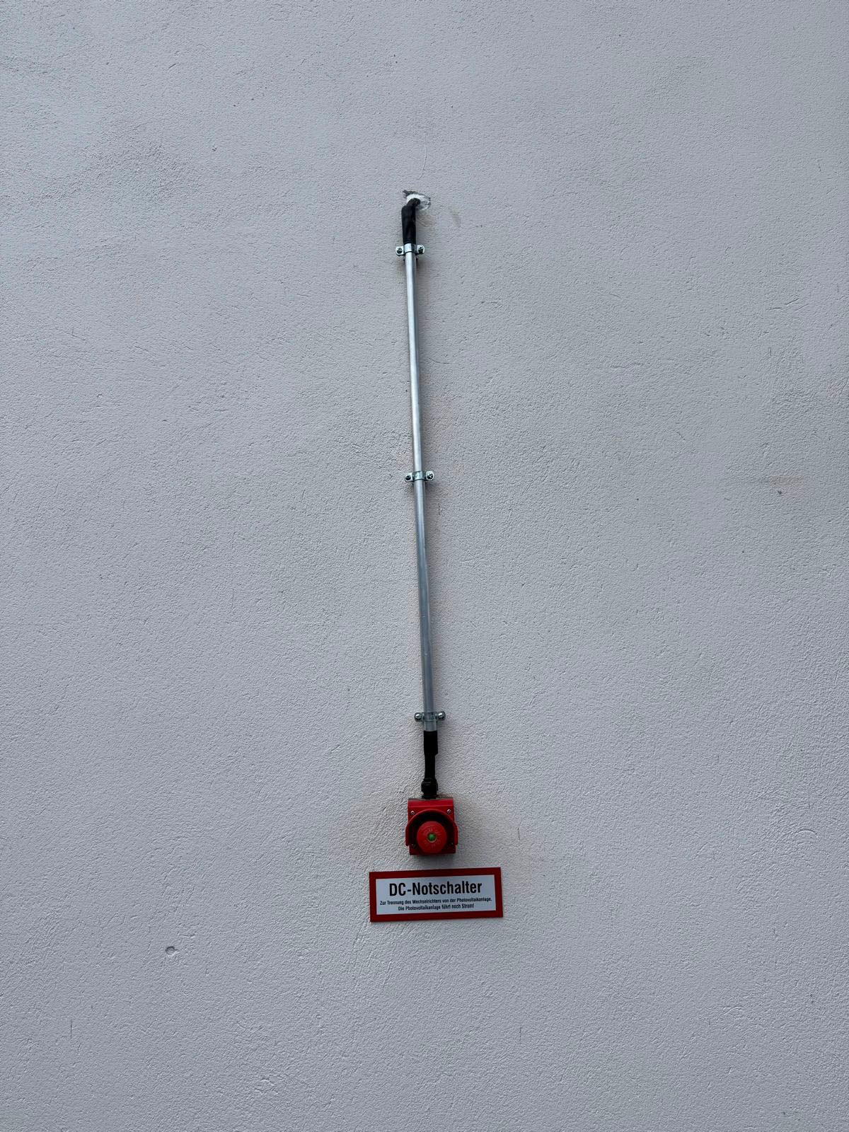 Roter Feuermelder mit Metallstange an einer weißen Wand.