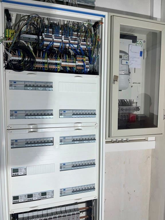 Elektrischer Verteilerkasten mit vielen Drähten und Sicherungsautomaten; rechts davon ein Schrank mit einer Tür.