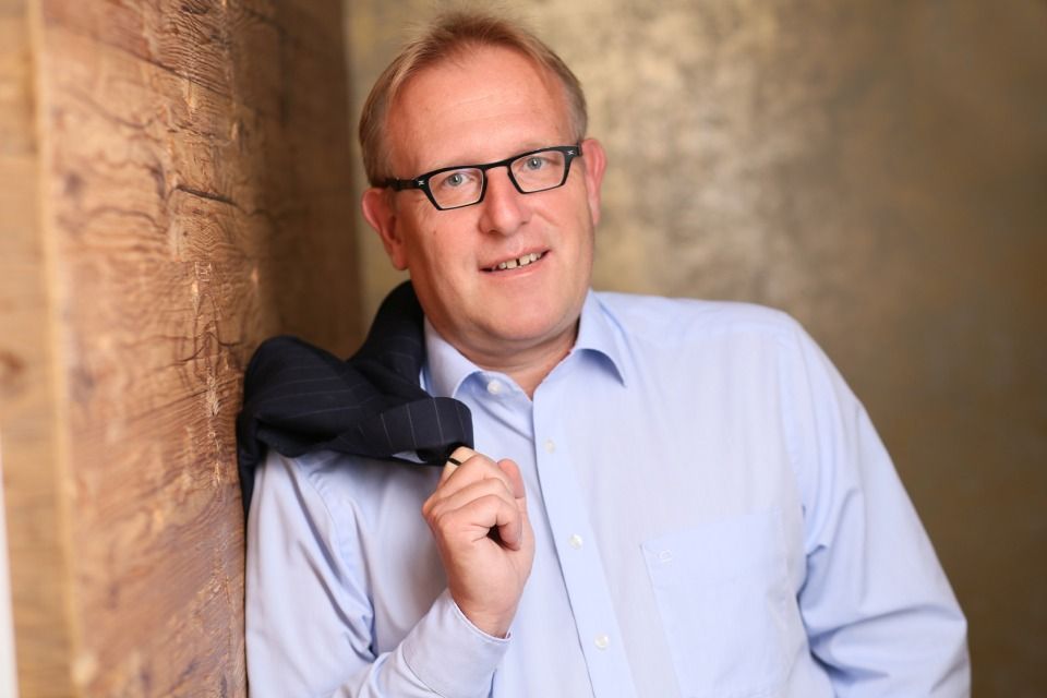 Rechtsanwalt Bernd Scheske - Ein Mann mit Brille und blauem Hemd lehnt an einer Holzwand.