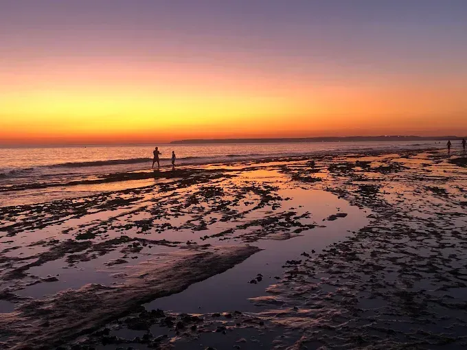 Puesta de sol sobre una playa rocosa con pozas de marea poco profundas y algunas personas cerca de la orilla.