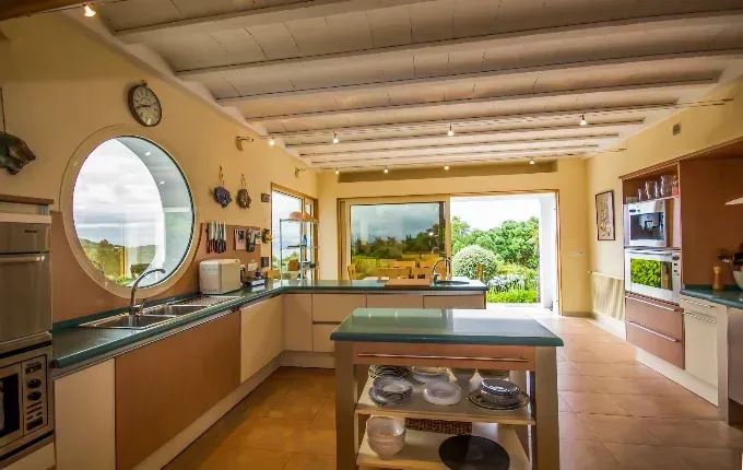 Cocina luminosa con armarios de madera, ventana redonda, isla y vistas despejadas a un jardín soleado.