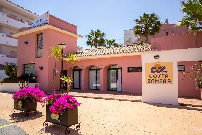 Entrada rosa al complejo turístico con palmeras, flores y un letrero que dice “C2 ZAHRAA”.