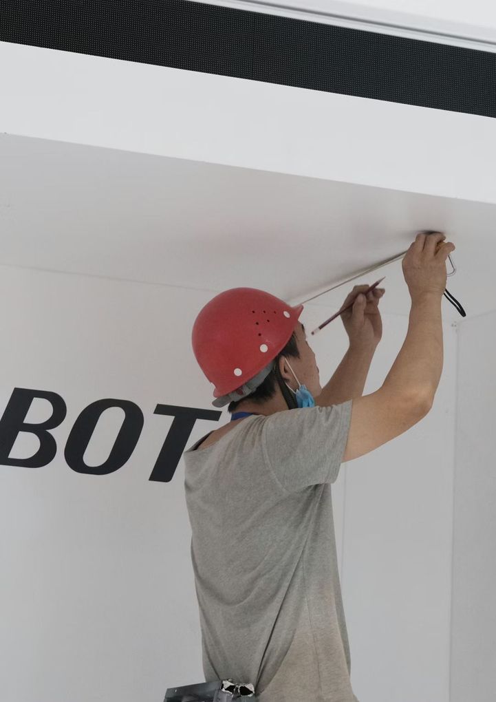 Une personne portant un casque de chantier rouge utilise un crayon et une règle pour tracer une ligne sur un plafond blanc, à côté d'un mur où figure du texte.