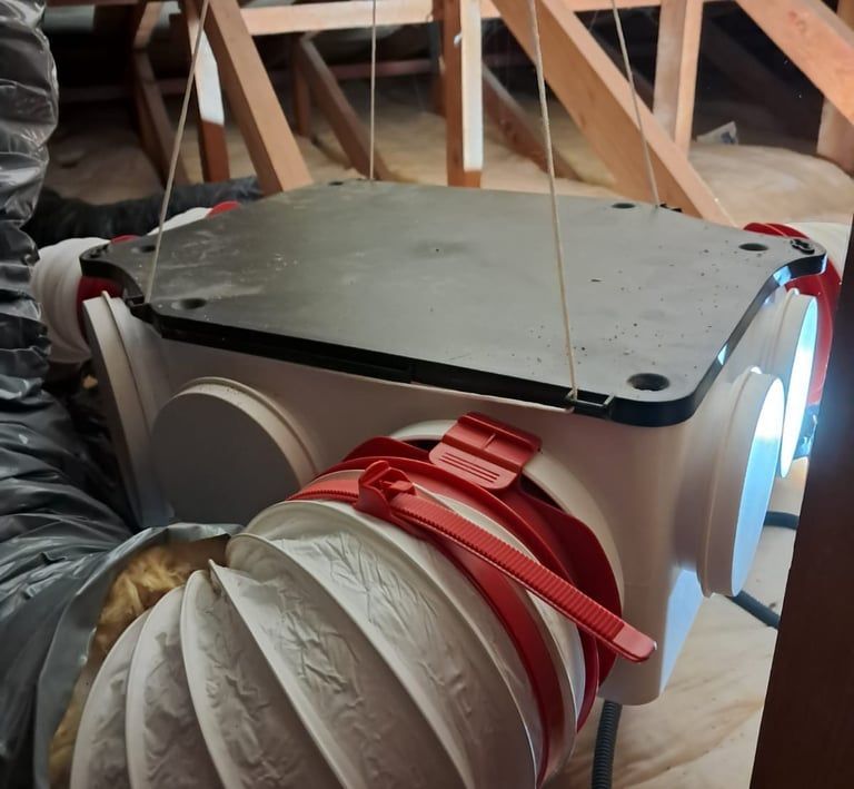 Un caisson de ventilation blanc avec un couvercle noir est suspendu dans un grenier et relié à une gaine flexible isolée par des attaches rouges.