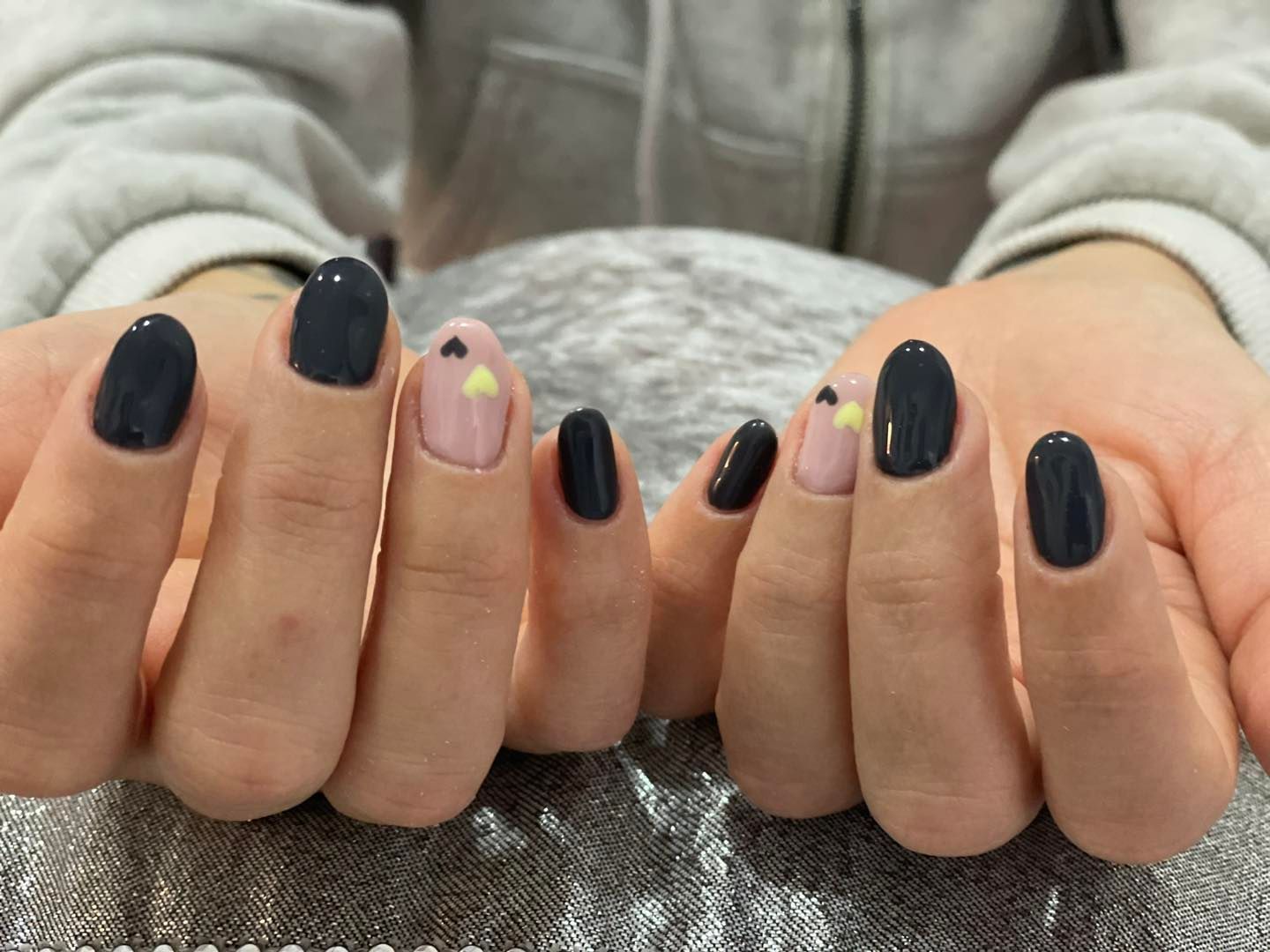 Manos con esmalte de uñas ovalado azul oscuro y rosa. Dos uñas tienen pequeños diseños de corazones amarillos y negros.